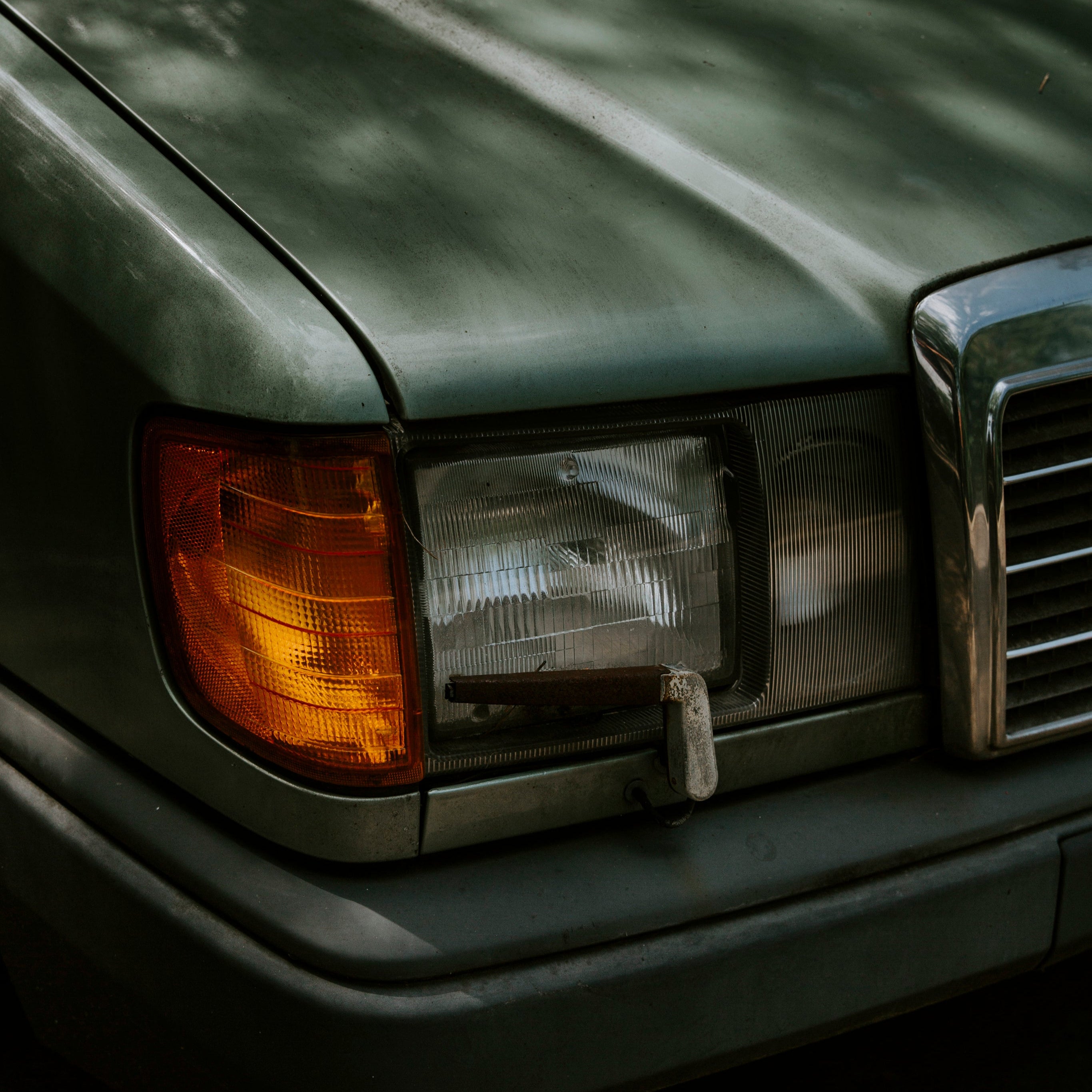 Nahaufnahme der Front eines klassischen dunkelgrünen Mercedes mit markantem Scheinwerfer und Chromdetails, fotografiert bei Tageslicht.
